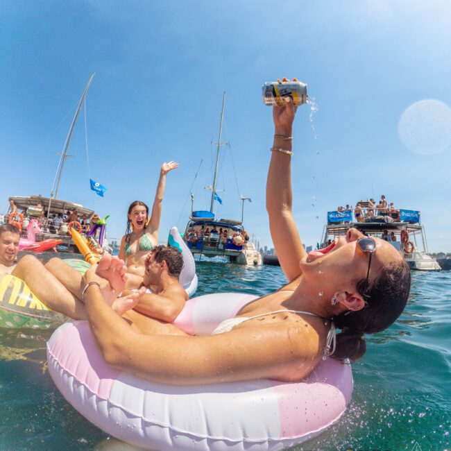 A woman in sunglasses lounges on a pool float in the sea, pouring a drink into her mouth, while others around her cheer and smile. Boats and people are visible in the background under a clear blue sky.
