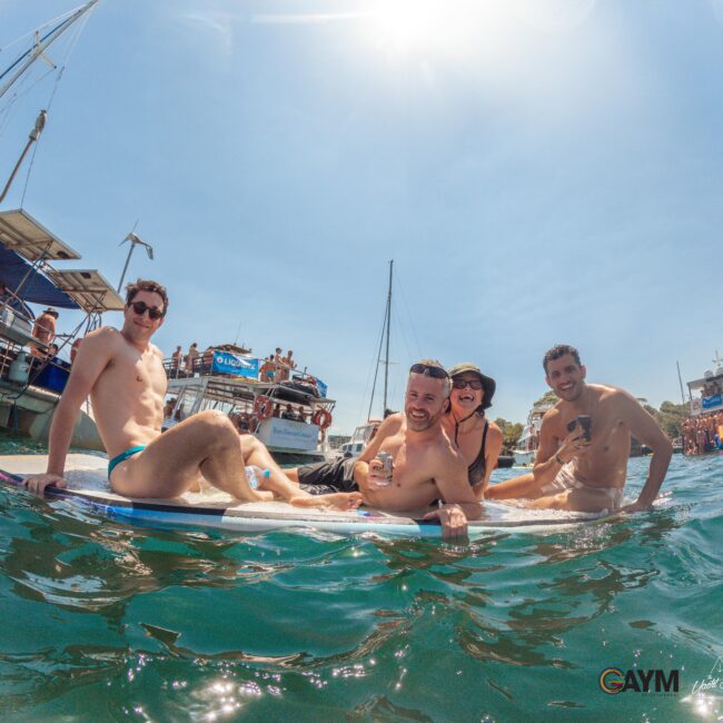 Four people in swimwear relax on a floating mat in the sea, smiling and holding drinks. Boats and a crowd are visible in the sunny background. The atmosphere appears festive and lively.