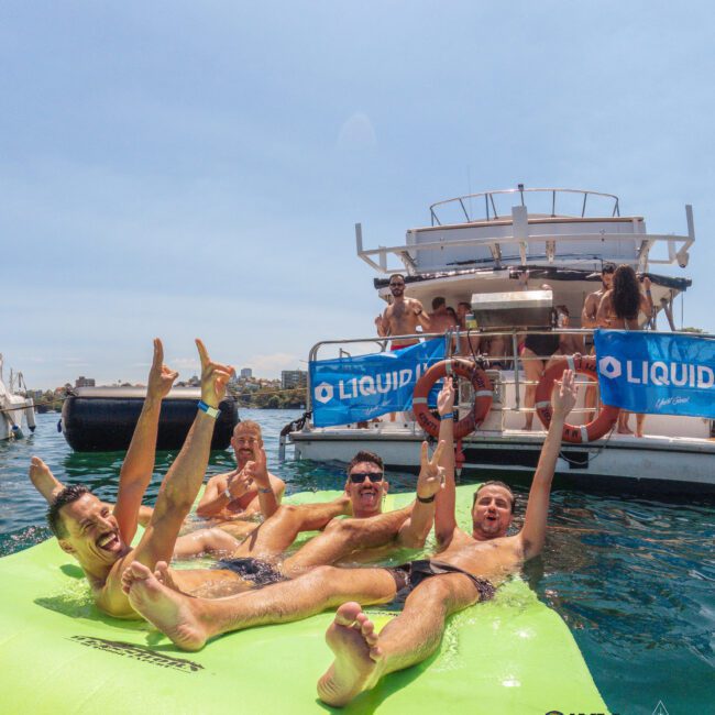 A group of smiling men in swimsuits relax and pose on a green floating mat in the water near a yacht, with other people visible on the boat under sunny skies. Blue "LIQUID" banners hang from the yacht.
