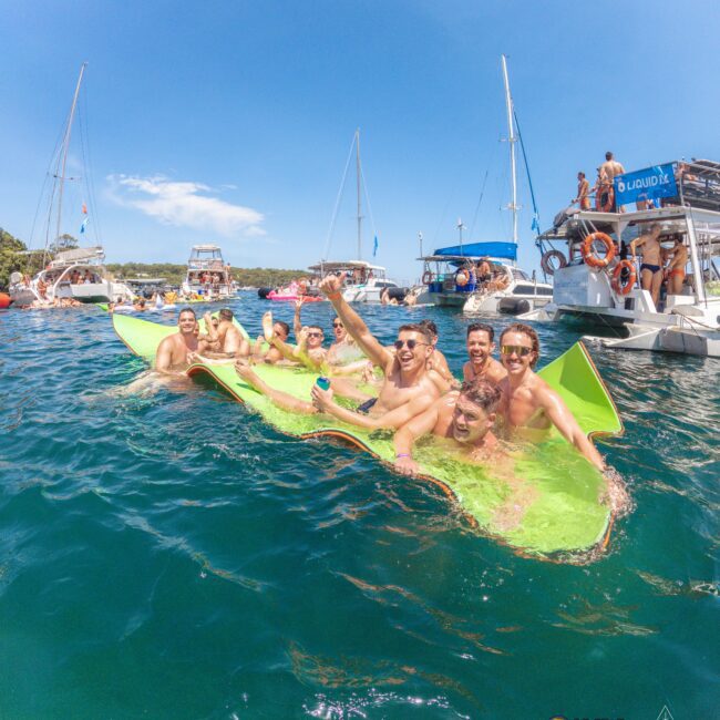 A group of smiling young men lounge on a floating mat in the water, surrounded by boats under a sunny blue sky at a lively summer party.