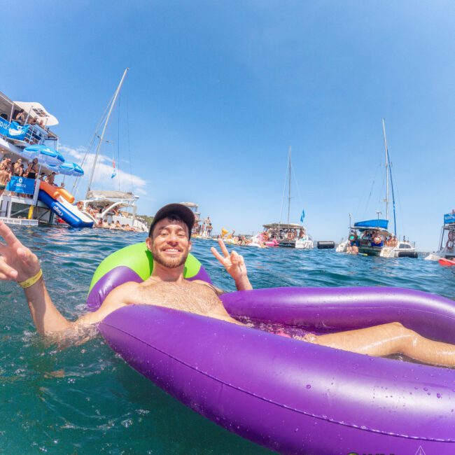 A man smiles and makes a peace sign while lounging on a purple inflatable in the ocean, surrounded by people, yachts, and boats under a clear blue sky.