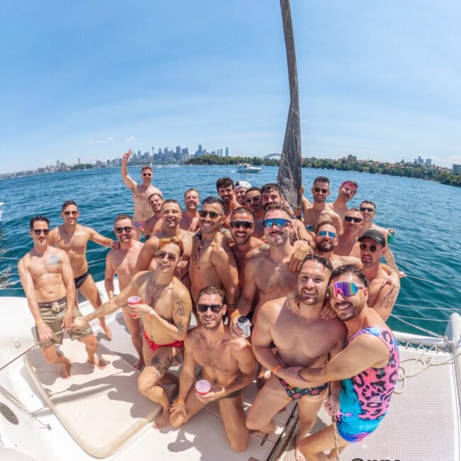 A group of men in swimwear smile and pose together on a boat under sunny skies, with a city skyline and water in the background. Some hold drinks, and everyone looks cheerful and relaxed.