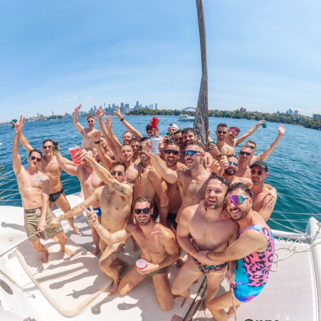 A group of men in swimwear smile, laugh, and raise their hands while posing together on a boat under sunny skies, with a city skyline and water in the background. The atmosphere is festive and joyful.