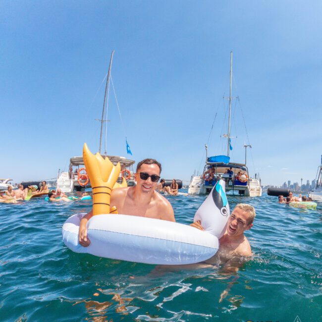 Two people smile and pose in the ocean near boats, holding an inflatable float shaped like a golden mermaid tail and a small inflatable orca. Blue skies and several boats with people in the background.