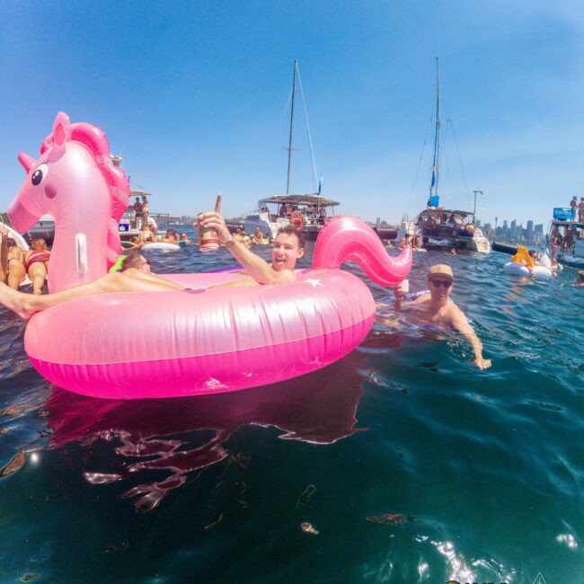 A person rides a large pink inflatable unicorn float in the water, surrounded by boats and other people swimming. The sky is clear and blue, and the city skyline is visible in the background.