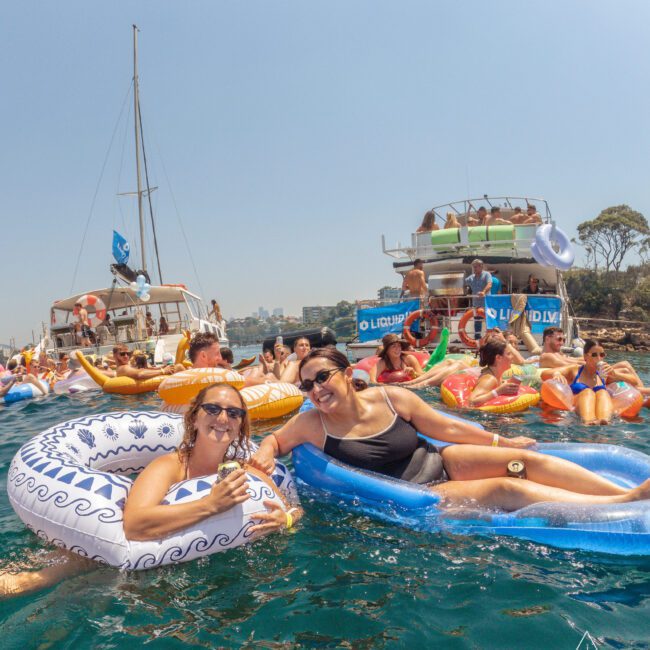 People relaxing on colorful inflatable rafts and pool floats in the water near anchored boats on a sunny day. Many are smiling and enjoying the festive atmosphere. Trees and blue sky are visible in the background.