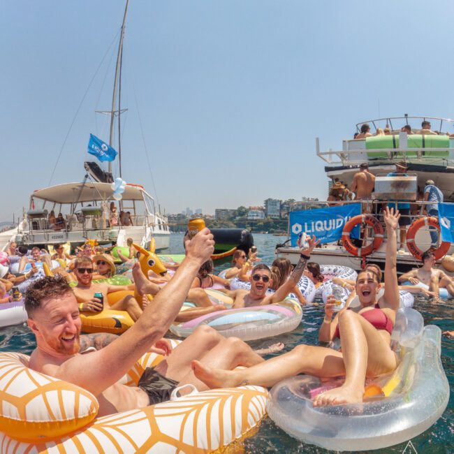 A group of people on inflatable floats enjoy drinks and celebrate together in the water near yachts on a sunny day, with city buildings visible in the background and a festive atmosphere.