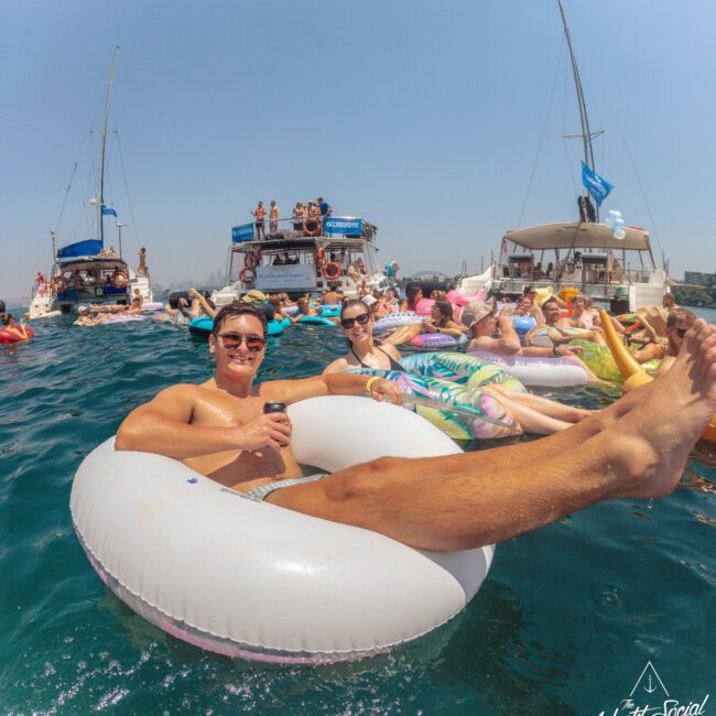 A group of people relax on colorful inflatable floats in the sea near several yachts. A man in sunglasses smiles at the camera while lounging on a white tube. The weather is sunny and festive.