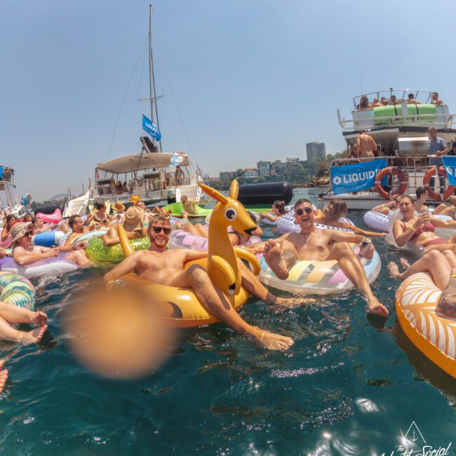 People relax on colorful pool floats in the water near boats during a sunny day. Some smile and pose for the camera, enjoying a festive and lively atmosphere. The Yacht Social Club logo is visible in the corner.