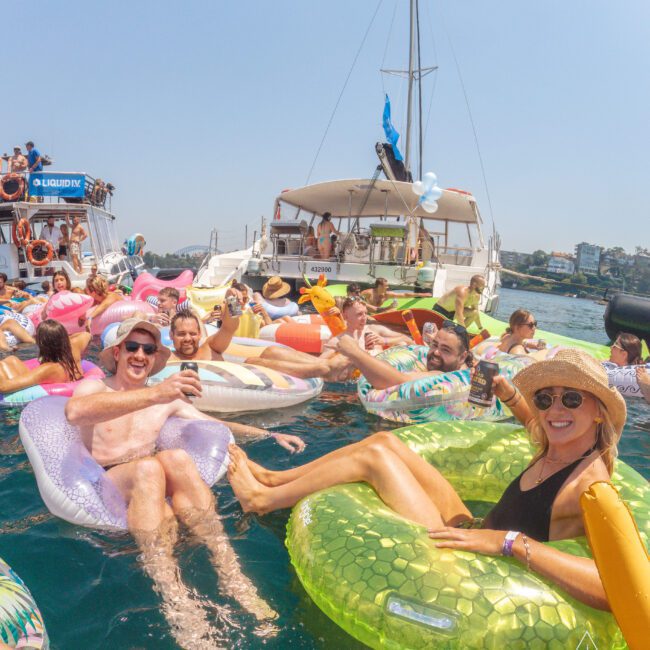 A group of people enjoy a lively party on inflatable floaties in the water near two docked boats, smiling, holding drinks, and wearing summer attire under a sunny, clear sky.