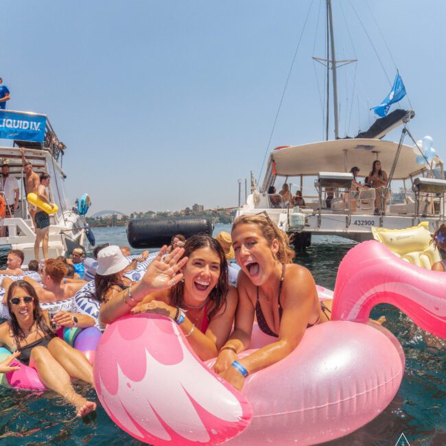 Two women smiling and waving while sitting on a large pink flamingo float in the water, surrounded by other people on floats near boats on a sunny day.