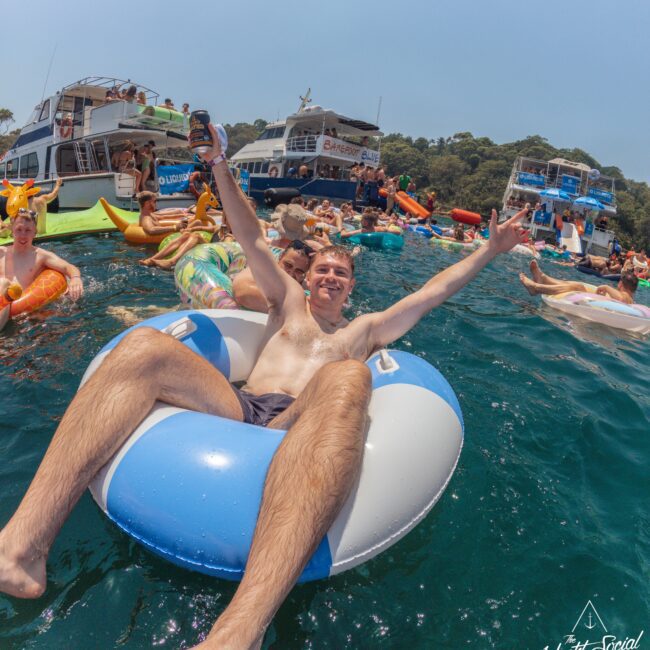 A man in swim trunks lounges on a blue and white inflatable ring, smiling and raising his arms in excitement while floating in the water among other people and boats during a lively summer party.