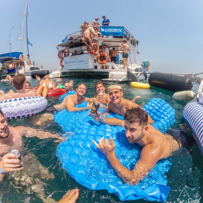A group of young people relax on blue pool floats and inflatable tubes in the water near a party boat, smiling and holding drinks under a clear sky. More people are gathered on the boat in the background.