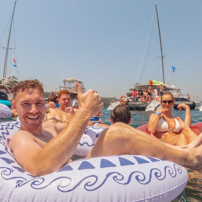 A man smiles and gives a thumbs up while lounging on an inflatable float in the water. Other people relax on floats nearby, with boats and a clear, sunny sky in the background.