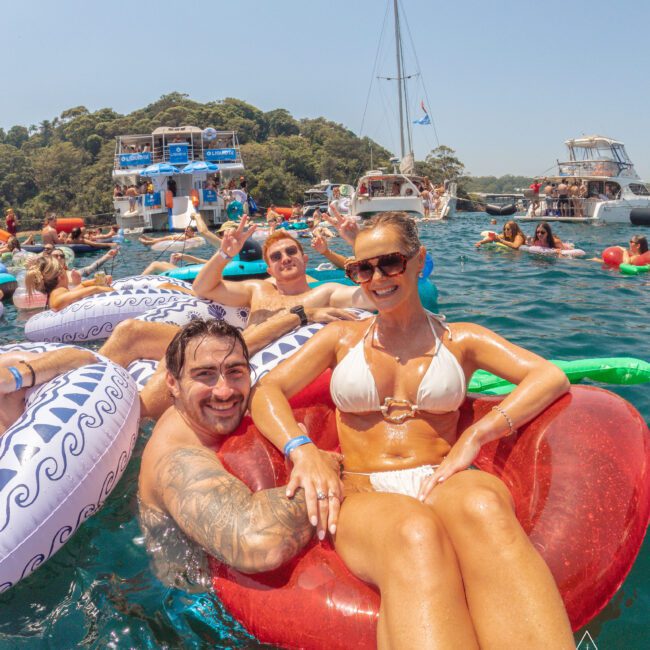 A group of people smiling and relaxing on colorful floaties in the water, with boats and more people in the background under a sunny sky. A woman in sunglasses sits on a red float in front.