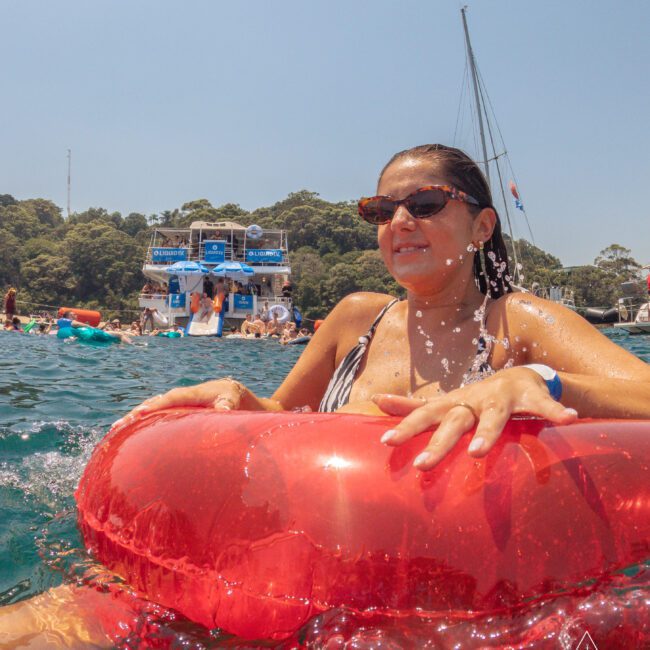 A woman in sunglasses floats on a red inflatable ring in the water, smiling. Behind her are other people, a boat, and a sailboat, with trees and a clear sky in the background.