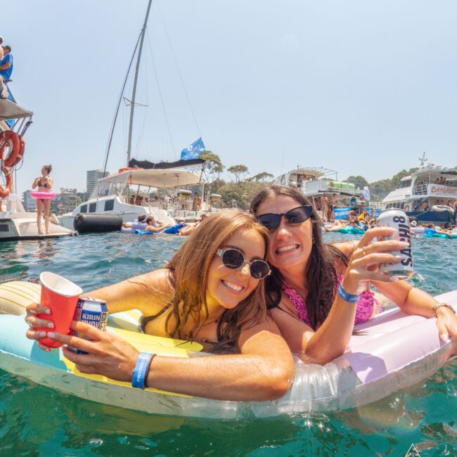 Two women in sunglasses smile and hold drinks while lounging on an inflatable float in a sunny, crowded party scene on the water, with boats and other people in the background.