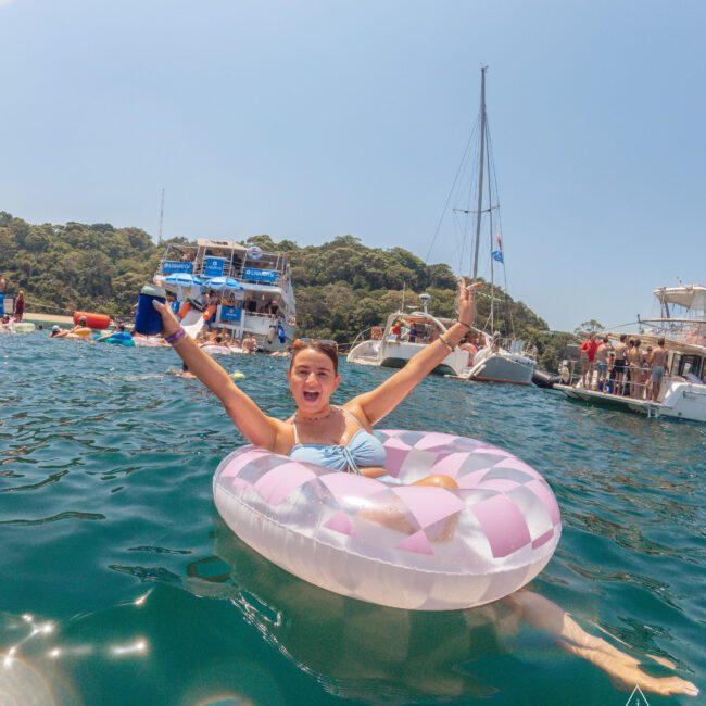 A woman in a pink and white inflatable ring floats in clear blue water, smiling and raising her arms. Boats and people are visible in the background, with green trees on the shore.