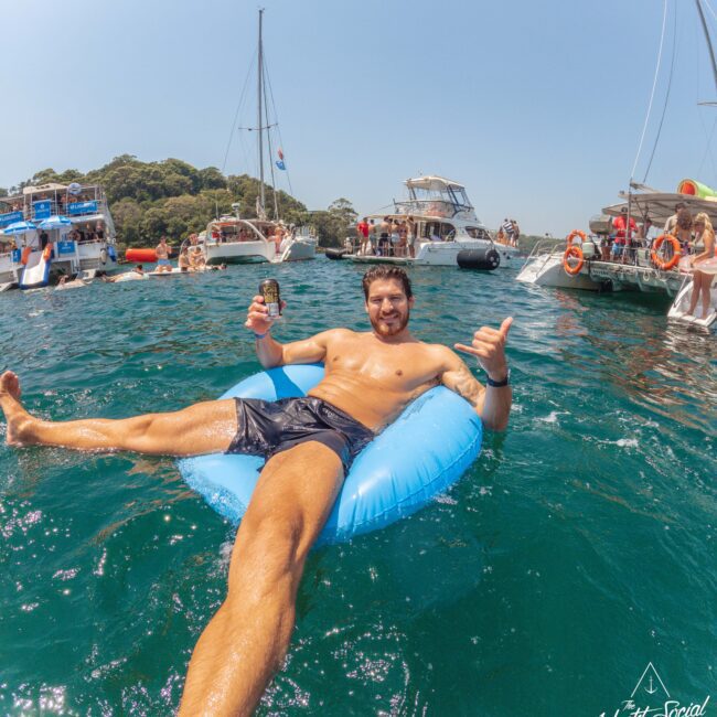 A man in swim trunks relaxes on a blue inflatable float in the water, smiling and giving a thumbs-up while holding a drink. Several yachts and people swimming are visible in the background under a sunny sky.
