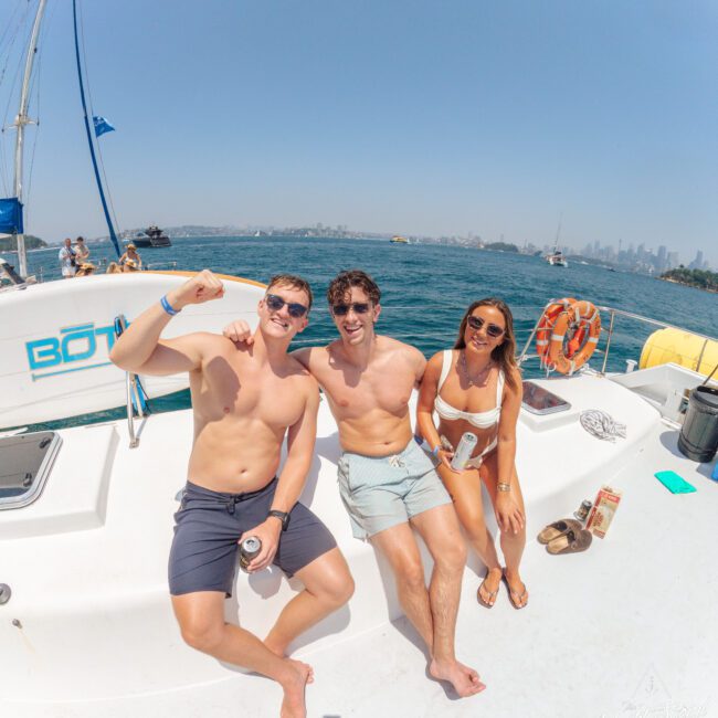 Three young adults in swimwear sit smiling on the deck of a boat under clear skies, with the ocean and a distant city skyline in the background. The atmosphere is relaxed and sunny.