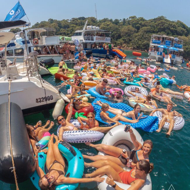 A large group of people in swimsuits relax on colorful inflatable floats between two boats in clear blue water, surrounded by forested hills under a sunny sky.