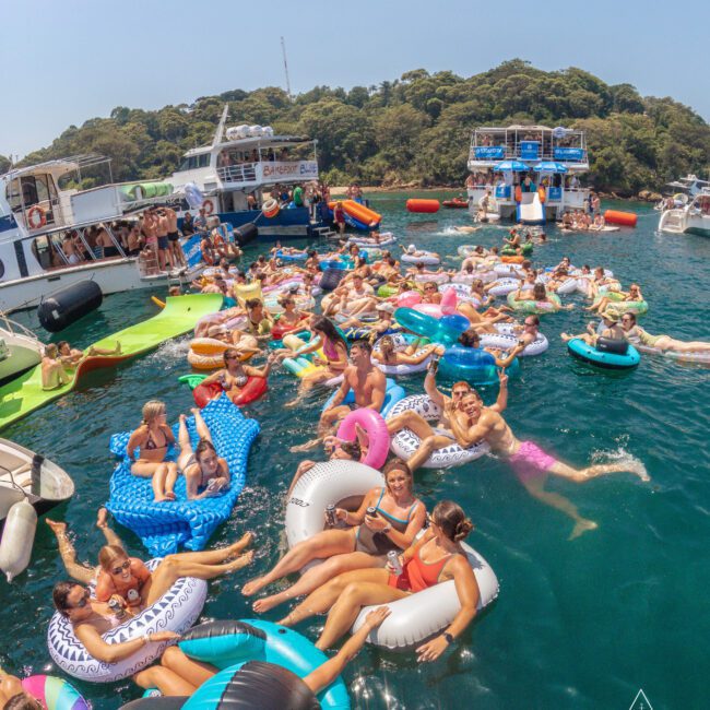 A large group of people relax and socialize on colorful inflatables in the water, surrounded by anchored boats on a sunny day, with lush trees in the background. The scene is lively and festive.