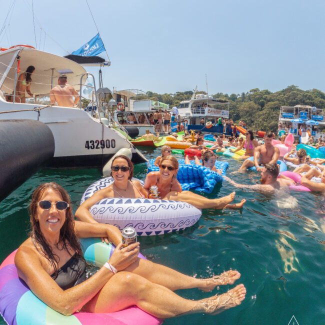 A group of people relax on colorful pool floats in the water near boats. Three women in sunglasses smile at the camera while holding drinks, surrounded by others enjoying the sunny day.
