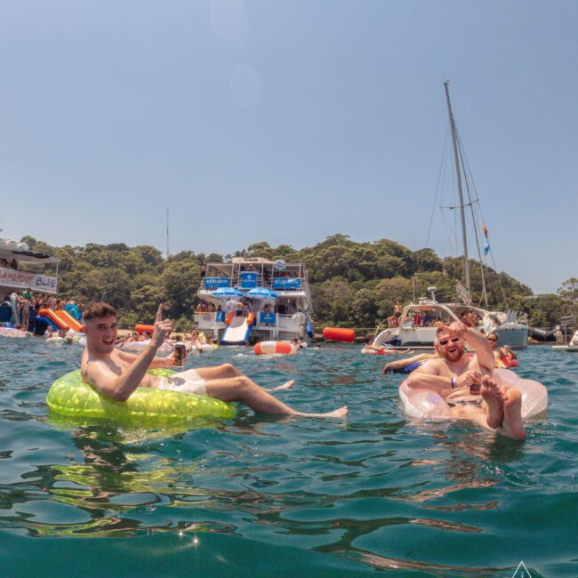 Two young men relax on inflatable pool floats in the water, smiling and making peace signs. Boats and more people are visible in the background, with trees and clear skies completing the sunny scene.