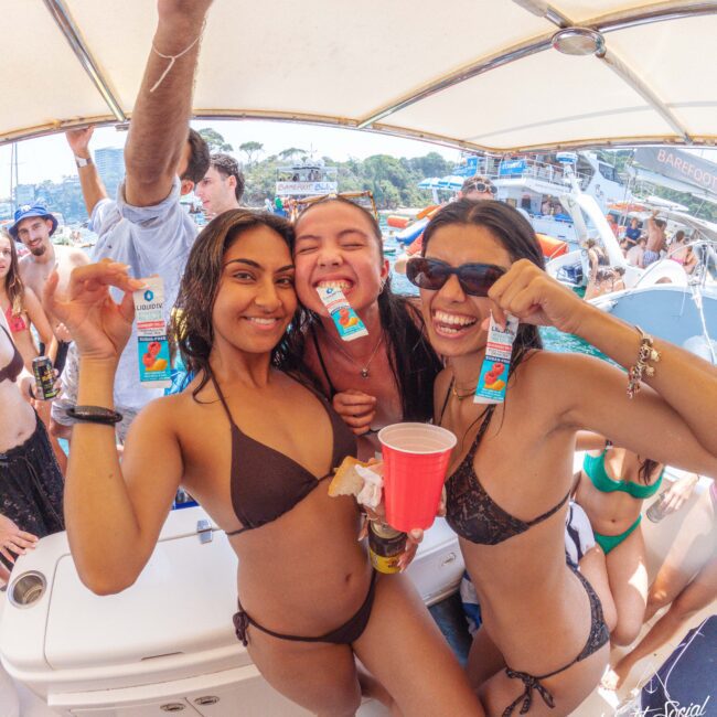 Three women in swimsuits smile and pose on a boat, holding colorful packets and drinks. Other people and boats can be seen in the background, suggesting a lively party atmosphere under a canopy.