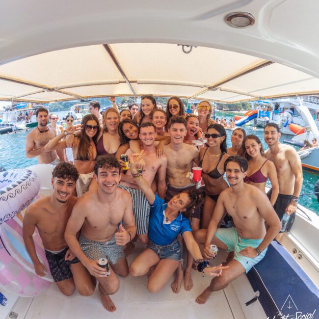 A large group of young adults in swimsuits smile and pose together on a boat, holding drinks, with other boats and people visible on the water in the background under a sunny sky.