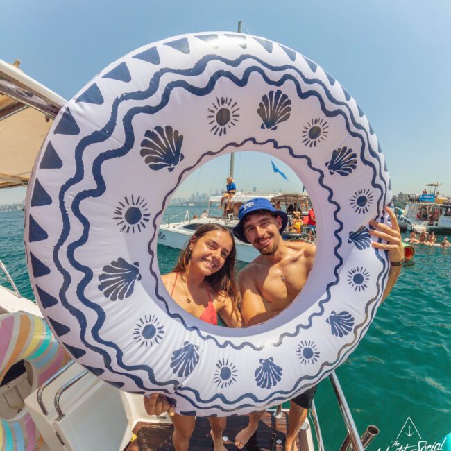 A smiling woman and man in swimwear pose on a boat, framed by a large inflatable pool ring with blue patterns. Other people and boats can be seen in the background on a sunny day.