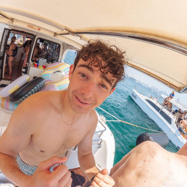 A young man with curly hair stands shirtless on a boat, smiling at the camera. People are socializing in the background, with inflatable floats and another boat visible on the water nearby. It’s a sunny day.
