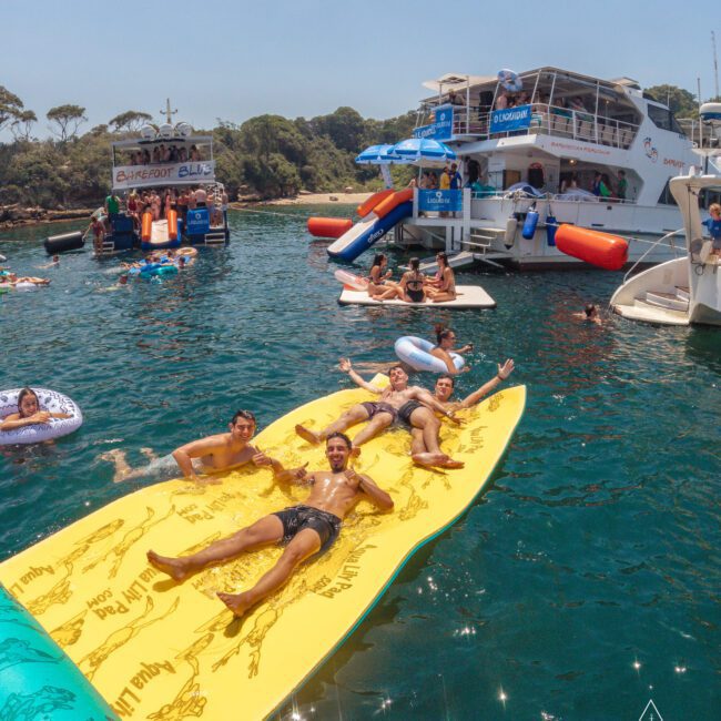 A group of people relax and pose on a large yellow floating mat in the water near boats. Others float on inflatables, and people are socializing around anchored yachts on a sunny day.