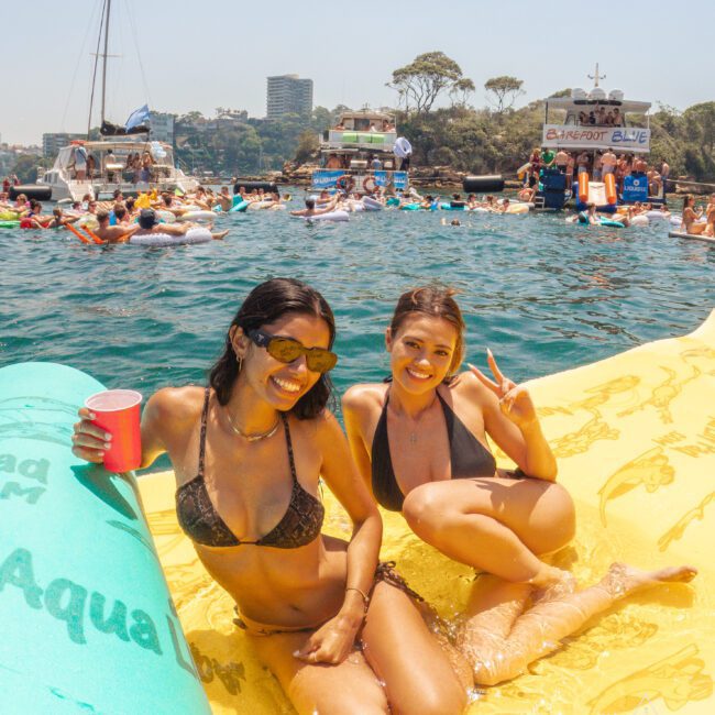 Two women in bikinis smile and pose on a large yellow float in the water, surrounded by other people, boats, and a lively summer party scene. One holds a pink cup, and the other makes a peace sign.