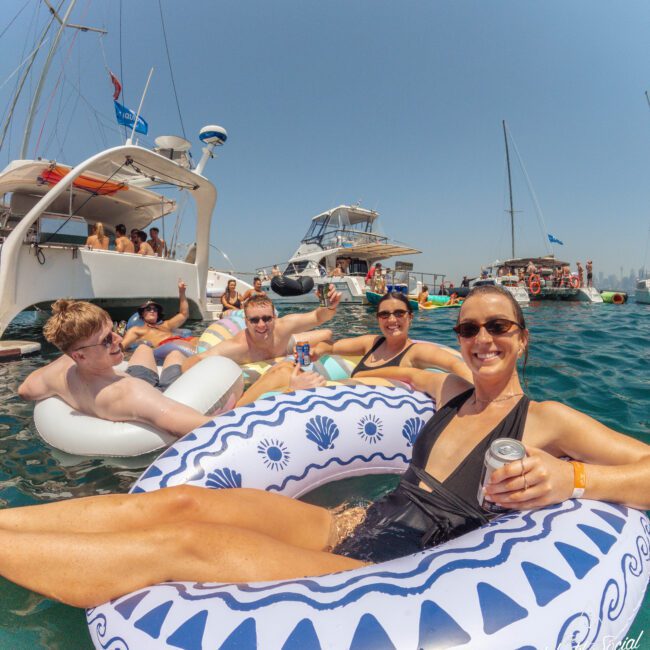 A group of people relax on inflatable pool floats in the water, smiling and holding drinks. Boats are anchored nearby under a clear blue sky, creating a fun and lively atmosphere.