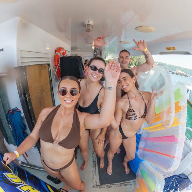 Four smiling people in swimsuits wave at the camera on a boat, surrounded by pool floats and water gear, with a scenic view of water and trees in the background.