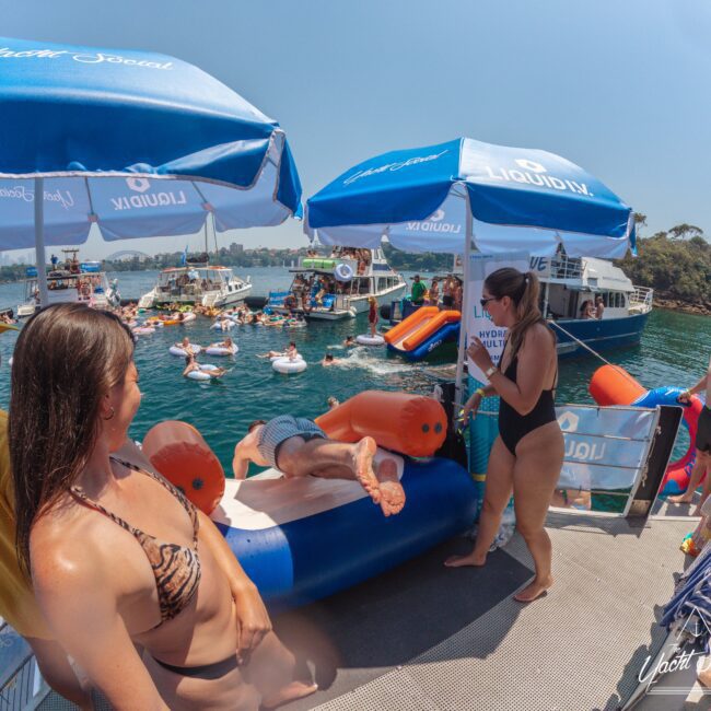 People relax and socialize on a boat deck with umbrellas by the water. Some are in swimsuits, others use inflatables, and several boats and floaties are seen in the background under a sunny sky.