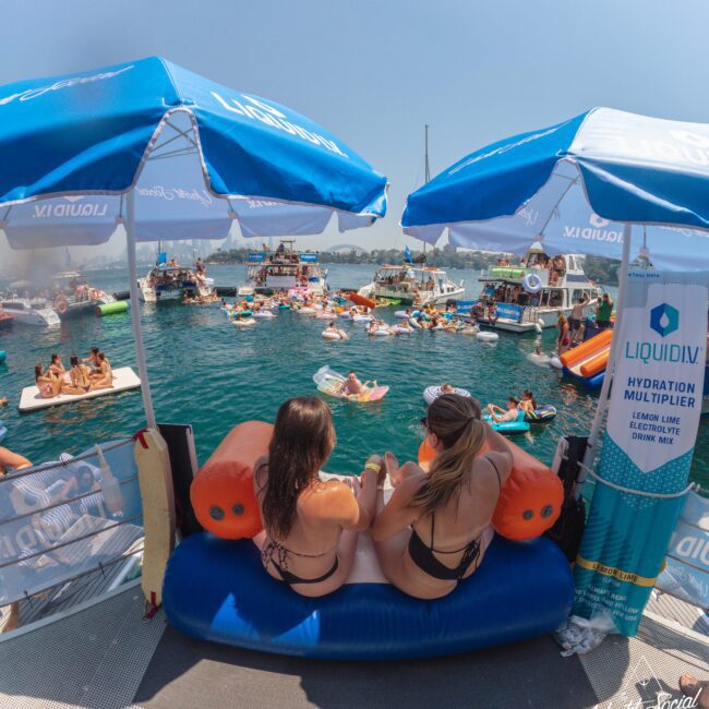 Two women in swimsuits sit on an inflatable couch under umbrellas on a dock, overlooking a crowded lake party with people on floats and boats. Liquid I.V. branding is displayed on the umbrellas and a nearby cooler.