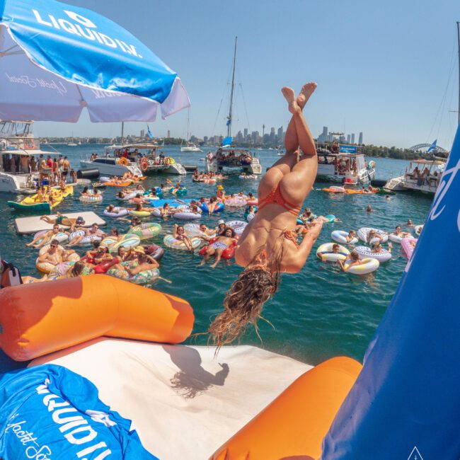 A woman in a bikini flips off an inflatable platform into the water, surrounded by people on colorful floats and boats, with a city skyline visible in the background on a sunny day.