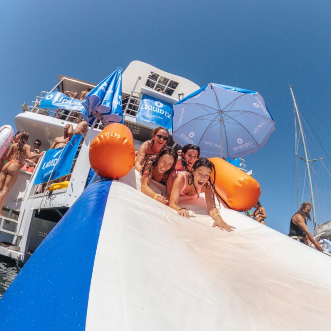 Four young people laugh and slide down a blue-and-white inflatable slide from a boat into the sea, with other people, umbrellas, and a yacht visible under a clear blue sky.