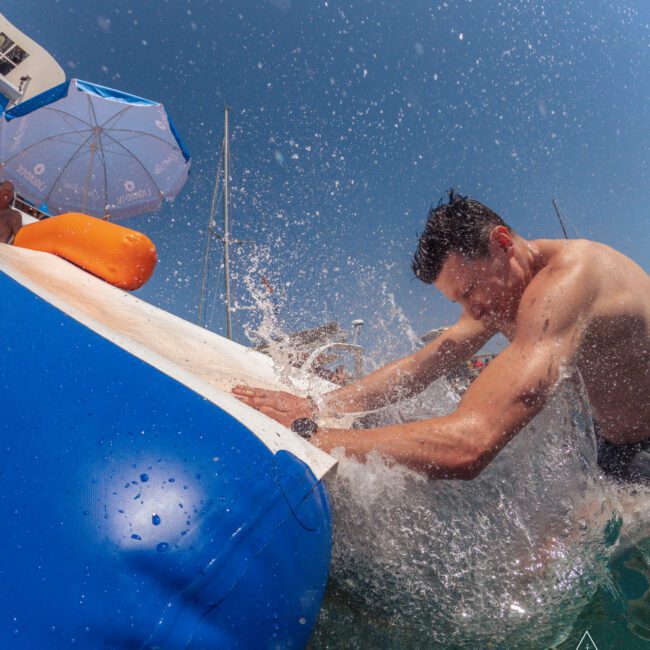 A man splashes into the water from a blue and white inflatable float under a clear sky, with an umbrella and boats visible in the background.