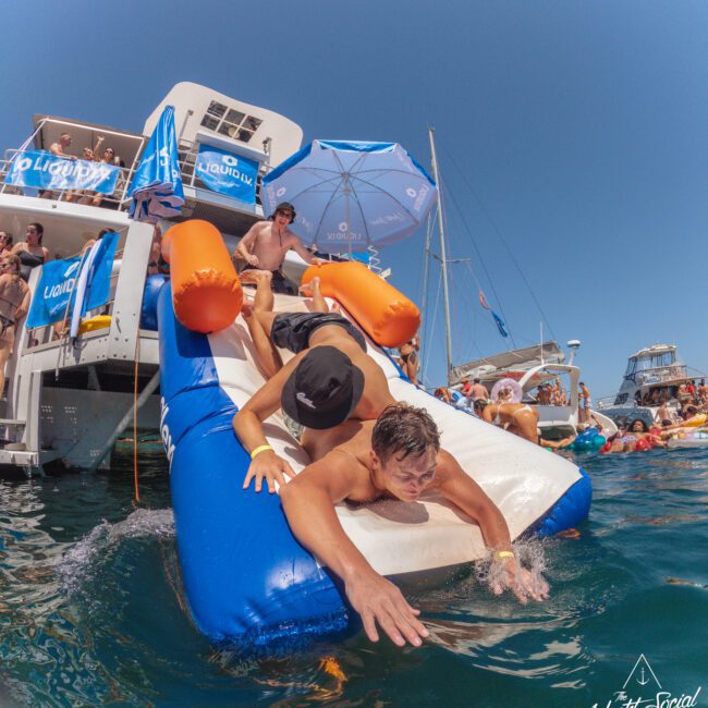 A group of people slide down an inflatable slide from a yacht into the ocean. It’s sunny, and the water is crowded with others enjoying the party atmosphere. A branded umbrella and logo are visible.