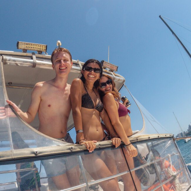 Three people in swimwear smile and pose together on a boat under clear blue skies, with other boats and the ocean visible in the background.