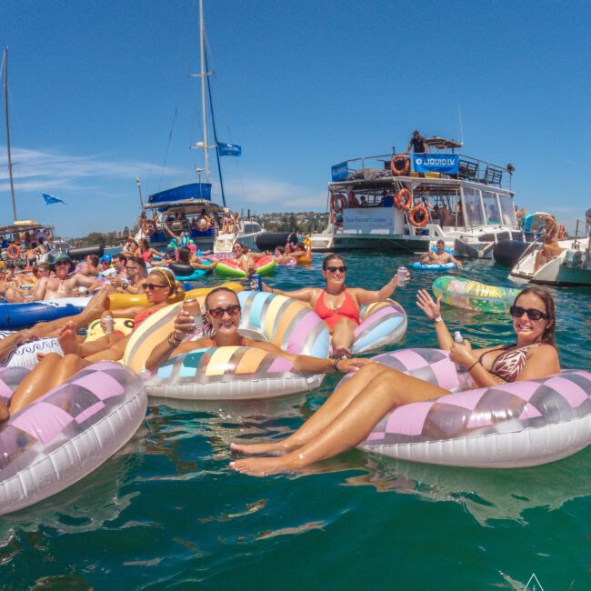 A group of people relax on colorful pool floats in the water near anchored boats, enjoying a sunny day. Others are on the boats and in the water, creating a lively, festive atmosphere.
