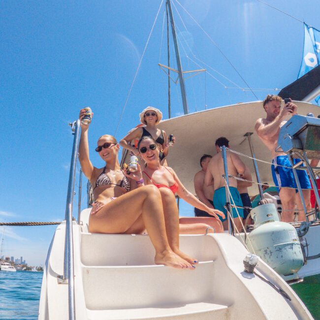 A group of people in swimwear relax and smile on a yacht's stairs, holding drinks. Blue sky and water are visible in the background, with more people enjoying the boat.