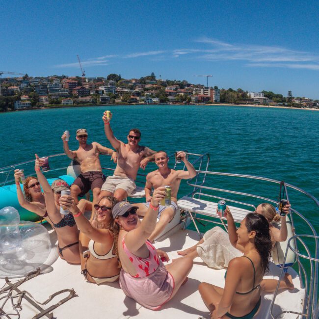 A group of people in swimsuits sit on a boat deck, smiling and raising drinks toward the camera on a sunny day, with blue water and a coastal town visible in the background.