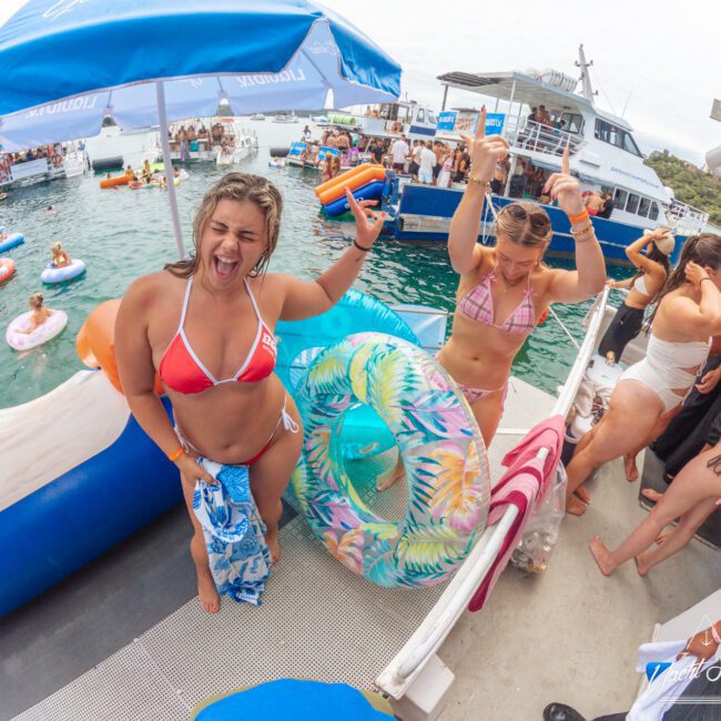 People in swimsuits dancing and laughing on a boat deck at a lively pool party, with others swimming and relaxing on floaties in the turquoise water and nearby boats under umbrellas in the background.