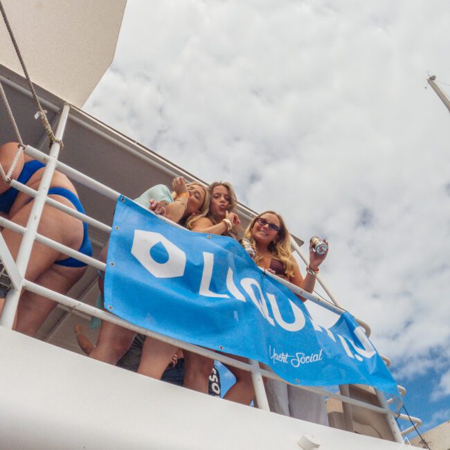 Several women in swimsuits stand on the deck of a boat, smiling and holding drinks, with a blue banner that reads "LIQUID" hanging on the railing. The sky is partly cloudy in the background.