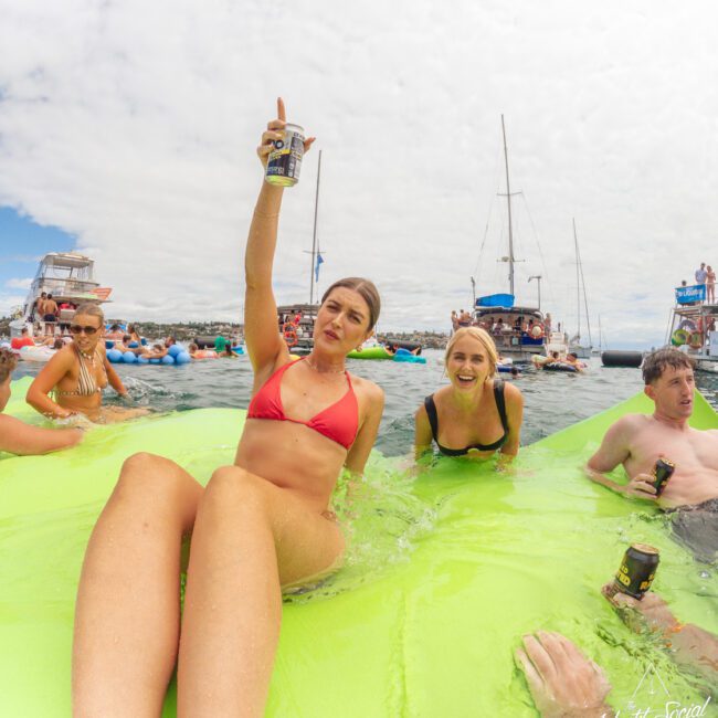 A young woman in a red bikini lounges on a bright green float, holding up a can with a smile, surrounded by friends in the water near several boats on a sunny day.