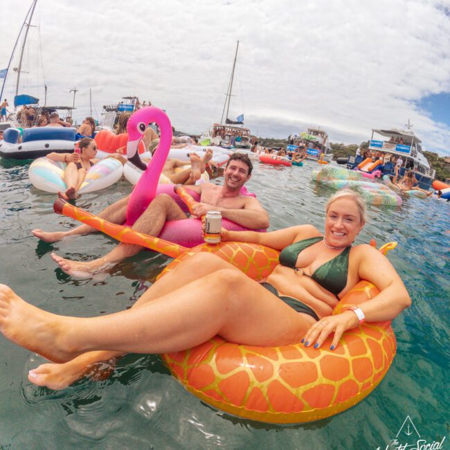 People relaxing on colorful inflatable floaties in the ocean, smiling and enjoying drinks. Boats are anchored nearby, and the sky is partly cloudy. The atmosphere is lively and festive.